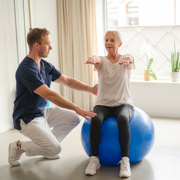Ältere Frau macht Übungen mit Physiotherapeuten auf einem blauen Gymnastikball und hält dabei ihre Arme nach vorne ausgestreckt.