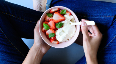 Eine Schale mit Erdbeeren und Joghurt in den Händen einer Person, die im Schneidersitz sitzt.