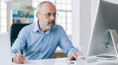 Ein älterer Mann im blauen Hemd und Brille arbeitet am Schreibtisch und blickt auf einen Computer-Bildschirm.