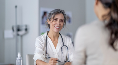 Ärztin mit Stethoskop lächelt und führt ein medizinisches Beratungsgespräch mit einer Patientin, die im Vordergrund sitzt.