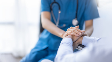 Nahaufnahme einer Ärztin in blauer Uniform mit Stethoskop, die die Hand eines Patienten hält.