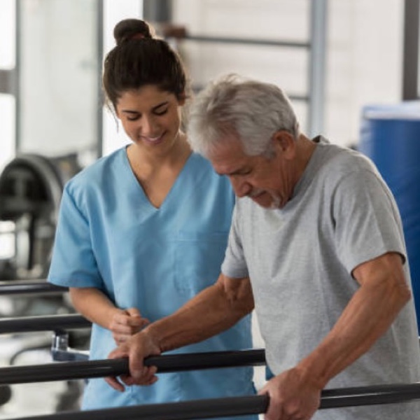 Älterer Mann wird von einer Physiotherapeutin in blauer Arbeitskleidung bei der Gehrehabilitation unterstützt.