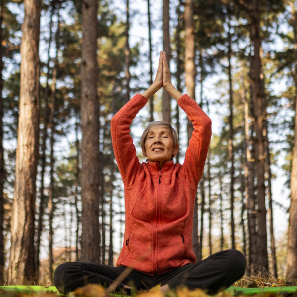Ältere Frau mit roter Jacke sitzt im Schneidersitz im Wald und macht Yoga.