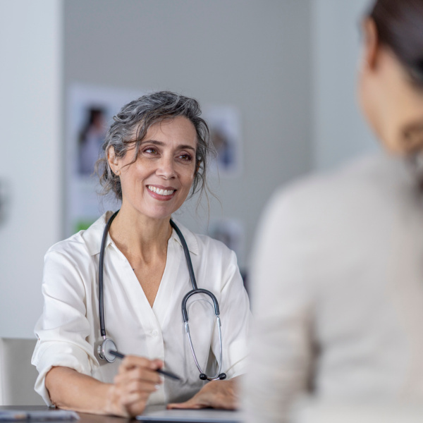 Ärztin mit Stethoskop lächelt und führt ein medizinisches Beratungsgespräch mit einer Patientin, die im Vordergrund sitzt.