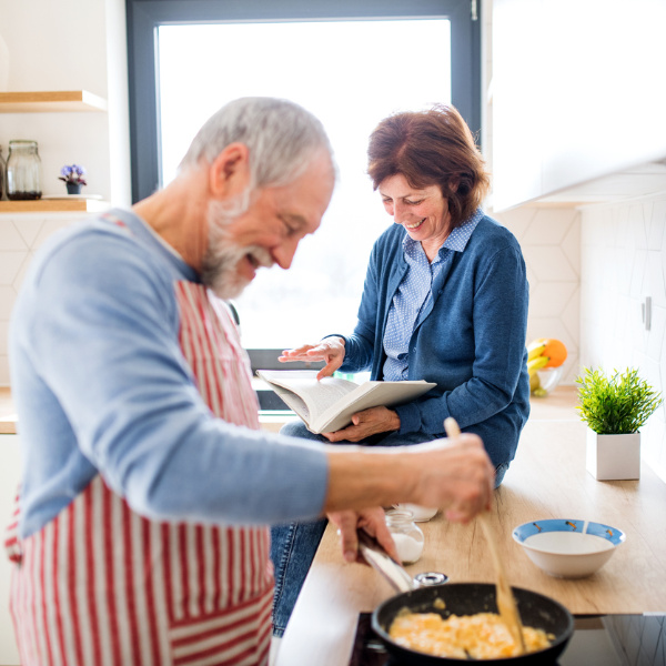 Ein älterer Mann mit gestreifter Schürze bereitet Essen in einer Pfanne zu, während die Frau auf der Küchenzeile sitzt und lächelnd durch ein Buch blättert.