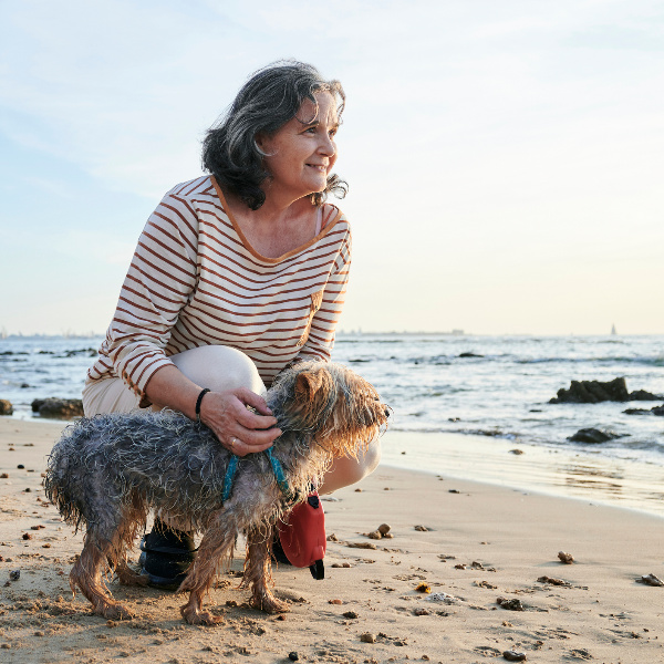 Eine ältere Frau hockt bei ihrem kleinen Hund am Strand und schaut Richtung Meer.