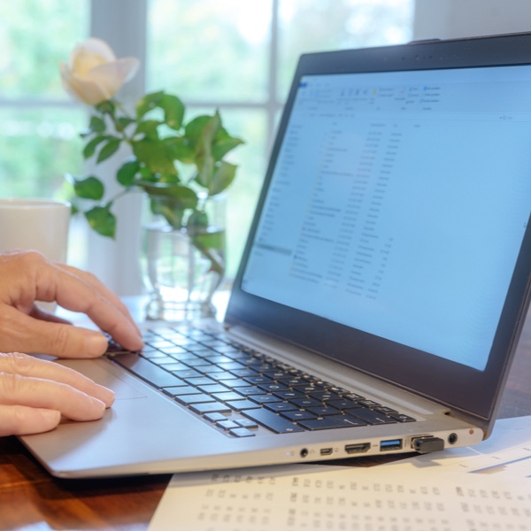 Hände einer älteren Person, die auf einem Laptop tippt. Im Hintergrund stehen eine Tasse und eine weiße Blume auf dem Schreibtisch.