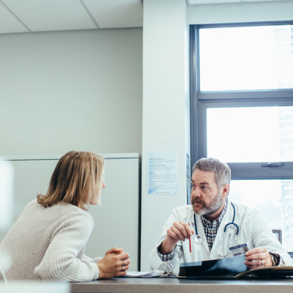 Ein Arzt mit Arztkittel und Stethoskop spricht mit einer Patientin am Schreibtisch im Büro.