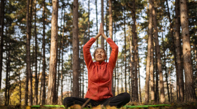 Ältere Frau mit roter Jacke sitzt im Schneidersitz im Wald und macht Yoga.